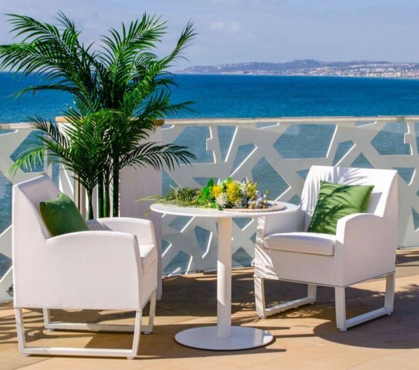 Two white armchairs in modern style with a round slim table in modern terrace with geometric lattice veranda and palm tree with a seafront background